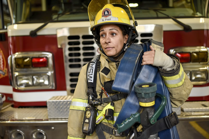 Female firefighter wearing full gear carrying a hose in front of a fire truck